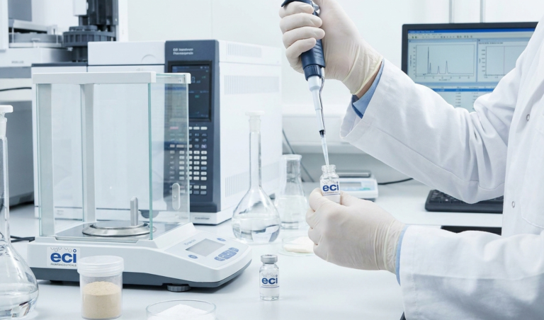 Lab technician in gloves using a micropipette to transfer liquid into a flask on a clean bench, with analytical instruments in the background.