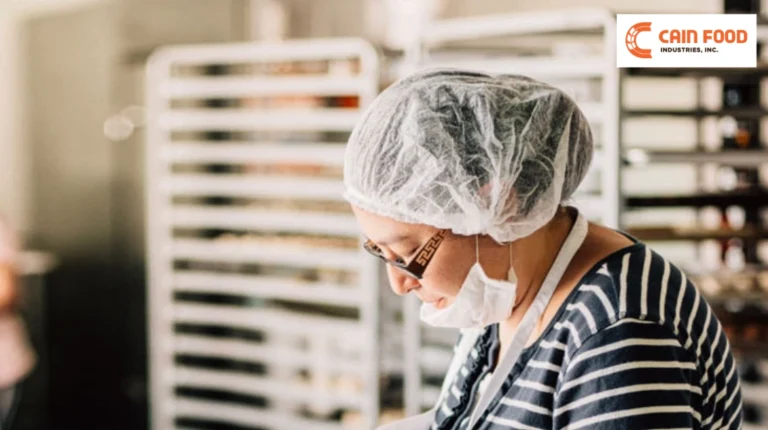 Bakery worker with hairnet and mask looking down in a kitchen with metal racks, Cain Food Industries logo in the corner.