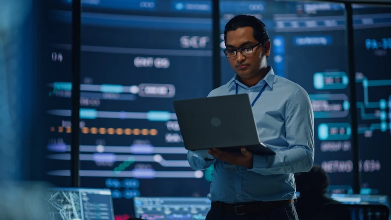 Man in a light blue shirt and glasses reviews data on a laptop in a high-tech control room with multiple screens displaying graphs and charts.