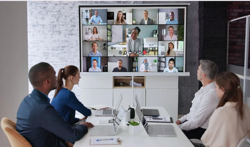 Team in a meeting room participates in a video conference shown on a large display with multiple remote participants.