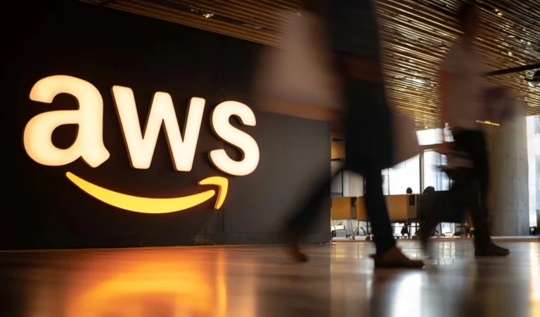 Bright AWS logo on a dark wall in a modern lobby; blurred people passing by in motion in the foreground.