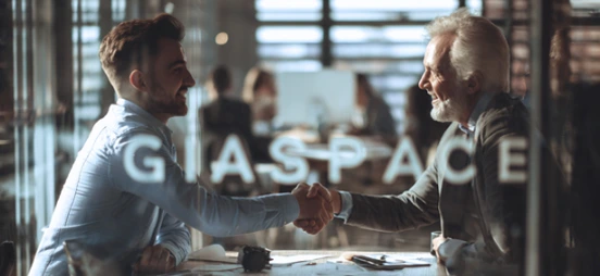 Two professionals shake hands across a conference table in a glass-walled office, with colleagues working in the background.